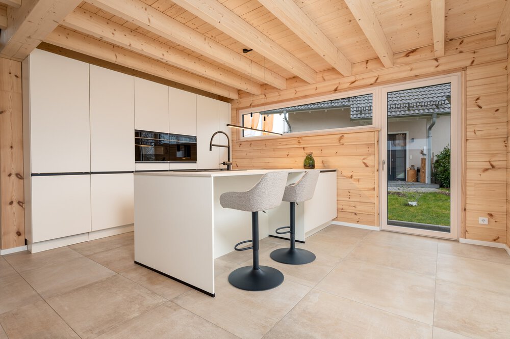 Moderne Küche mit Holzdecke und -wänden, cremefarbenen Schränken, Kochinsel mit Barhockern und großer Glastür zum Garten.