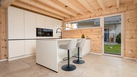 Moderne Küche mit Holzdecke und -wänden, cremefarbenen Schränken, Kochinsel mit Barhockern und großer Glastür zum Garten.