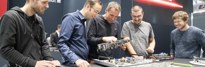 Fünf Männer stehen an einem Stand auf der productronica und betrachten begeistert elektronische Komponenten die auf einem großen Tisch ausgestellt sind.