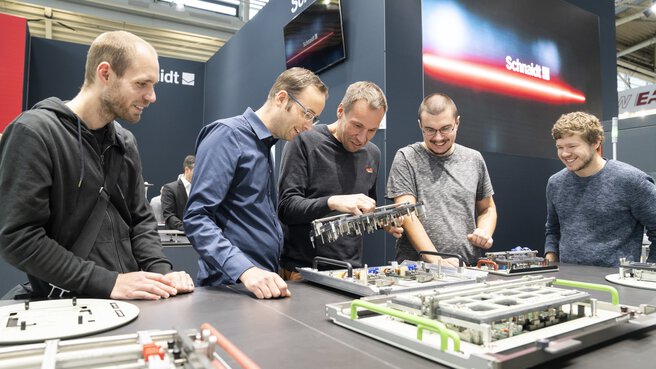 Fünf Männer stehen an einem Stand auf der productronica und betrachten begeistert elektronische Komponenten die auf einem großen Tisch ausgestellt sind.