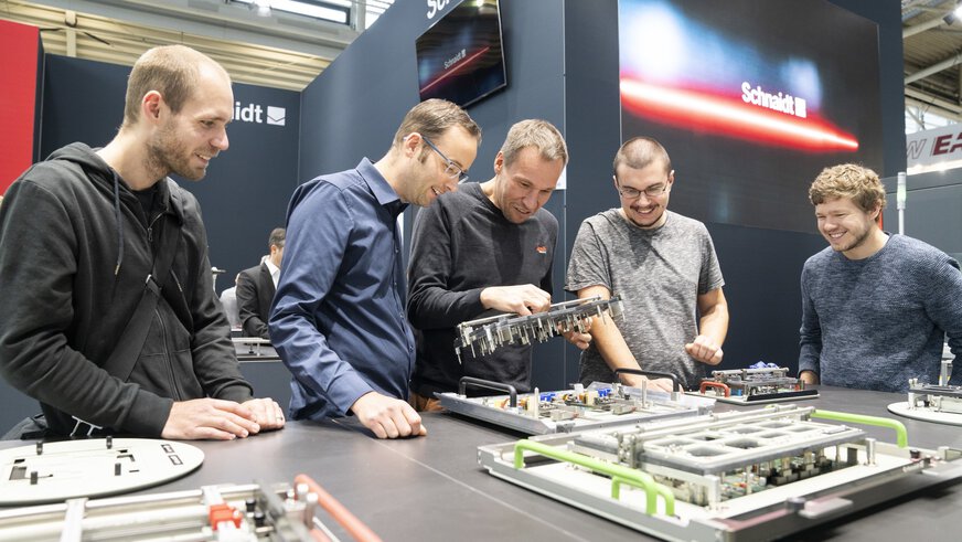 Fünf Männer stehen an einem Stand auf der productronica und betrachten begeistert elektronische Komponenten auf einem großen Tisch.