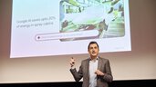 A speaker in a gray suit holds up a remote control. Behind him, a slide is displayed about saving energy with the help of Google AI.