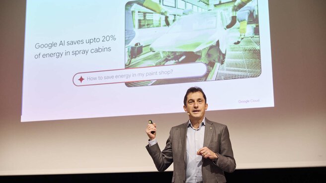 A speaker in a gray suit holds up a remote control. Behind him, a slide is displayed about saving energy with the help of Google AI.