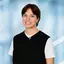 Smiling person with short hair, wearing a black vest over a white shirt, standing in front of a blue abstract background.