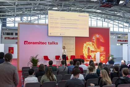 Eine Rednerin auf der Bühne der ceramitec talks spricht vor einem Publikum. Auf der Leinwand über Ihr wird eine Agenda angezeigt.