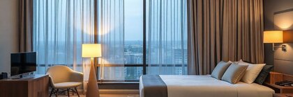 A hotel room with two double beds, a small television, chair, lamps, curtains and a window front offering a view over a city.