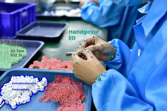 Eine Person in Laborkleidung demonstriert eine Handgeste. Vor ihr auf dem Tisch liegen Tabletts mit Kleinteilen, die farblich unterschiedlich markiert sind.