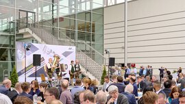 Menschen sitzen an Tischen im Freien mit einer Bühne und Musikern vor einer Messehalle.