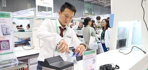 A man demonstrates laboratory equipment at a busy exhibition stand; visitors can be seen in the background.