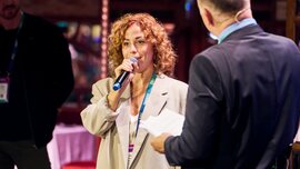 Eine Frau mit Konferenzbadge spricht mit einem Mikrofon auf einer Bühne und wendet sich einem Moderator im Anzug zu, der Notizen in der Hand hält; im Hintergrund sind ein Kronleuchter und die Einrichtung eines Veranstaltungssaals zu sehen.