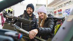 Zwei Personen stehen an einem Messestand für Fahrräder und schauen sich ein Fahrrad genauer an. Beide Personen tragen Mützen und Jacken in verschiedenen Farben.