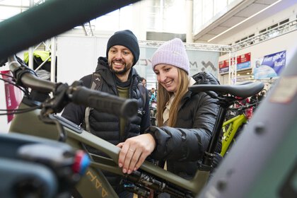 Two people are standing at a trade fair stand for bicycles and taking a closer look at a bicycle. Both people are wearing caps and jackets in different colors.