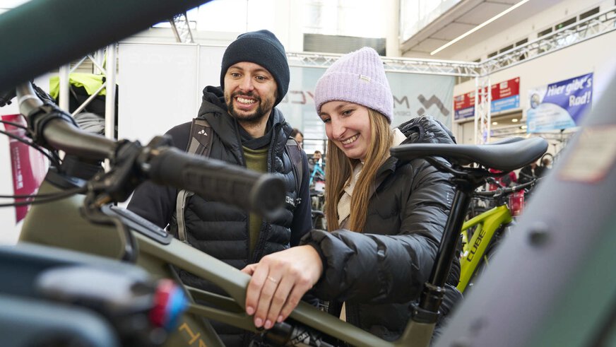 Zwei Personen stehen an einem Messestand für Fahrräder und schauen sich ein Fahrrad genauer an. Beide Personen tragen Mützen und Jacken in verschiedenen Farben.