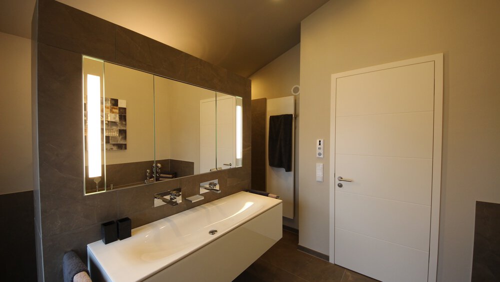 Blick in ein eingerichtetes modernes Badezimmer mit extra breitem Waschbecken mit zwei Wasserhähnen und großer Spiegelfront.