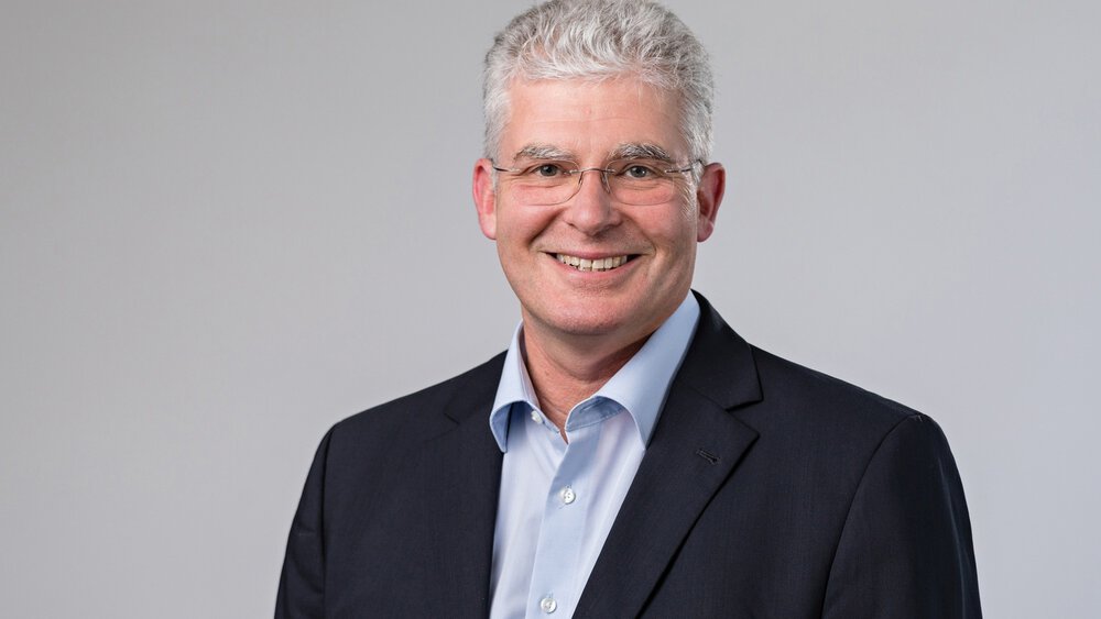 A smiling man with short grey hair and glasses, wearing a light blue shirt and black jacket, standing in front of a grey background.