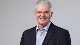 A smiling man with short grey hair and glasses, wearing a light blue shirt and black jacket, standing in front of a grey background.
