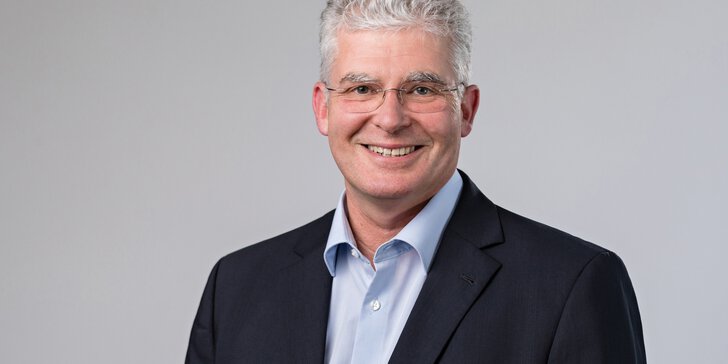 A smiling man with short grey hair and glasses, wearing a light blue shirt and black jacket, standing in front of a grey background.