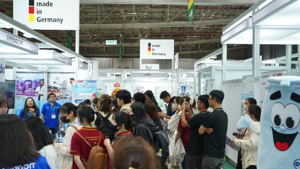Crowd at a trade fair with “Made in Germany” signs and a mascot on the right.