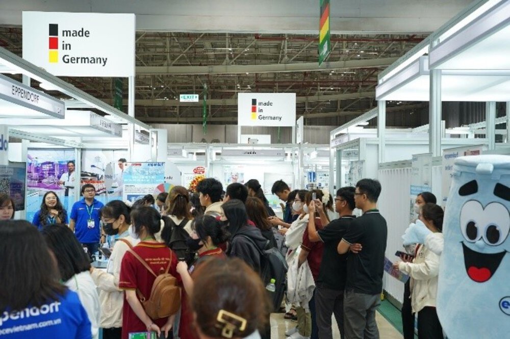 Crowd at a trade fair with “Made in Germany” signs and a mascot on the right.