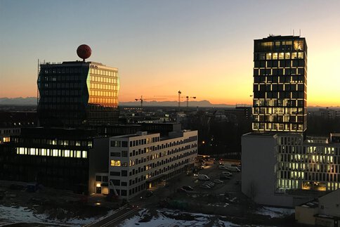 Blick aus der Vogelperspektive auf zwei große moderne Gebäude, Parkplätze und Baukräne im Abendrot.
