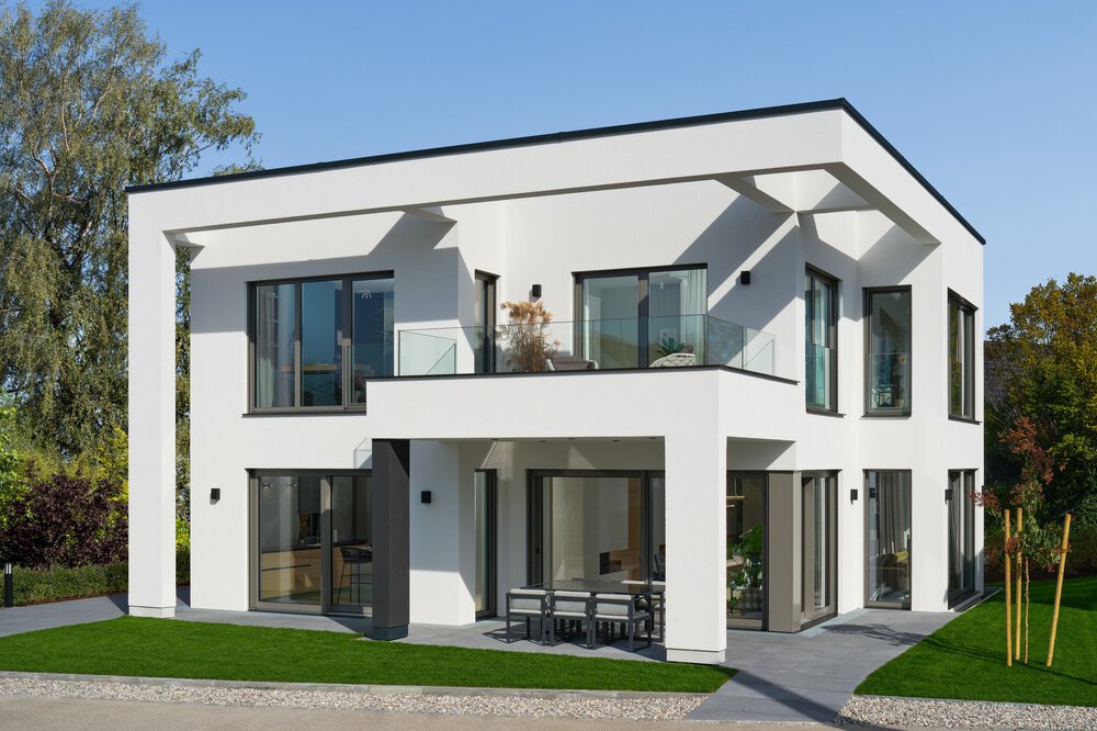 Modernes zweistöckiges weißes Haus mit großen Fenstern, einem Balkon und umgeben von Grün.