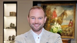 A smiling man with short hair and a full beard has his arms crossed and is wearing a watch, several rings, and a gray checkered suit.