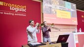 Zwei Männer mit Blasinstrumenten stehen mit einem Laptop und einem Soundboard auf einer Bühne der transport logistic und geben eine Vorstellung.