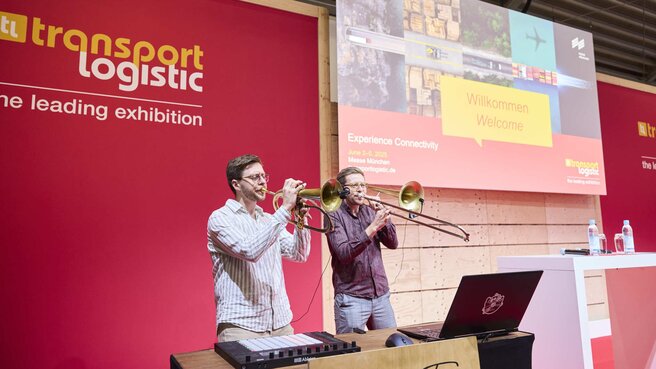 Zwei Männer mit Blasinstrumenten stehen mit einem Laptop und einem Soundboard auf einer Bühne der transport logistic und geben eine Vorstellung.
