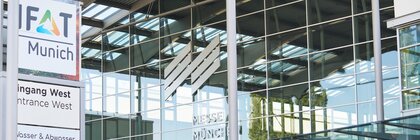 A close-up view of the western entrance to Messe München during IFAT, showing the reflective glass façade and the Messe München logo.