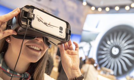 Smiling woman with a colorful scarf looks through a virtual reality headset at a trade fair stand.