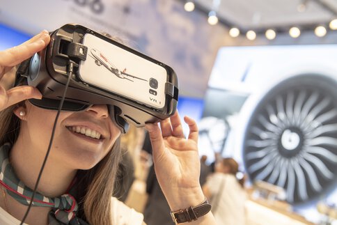 Smiling woman with a colorful scarf looks through a virtual reality headset at a trade fair stand.