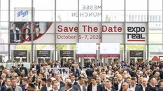 A large crowd of people attends the real estate trade fair EXPO REAL in the entrance hall of Messe München. Above the visitors hangs a large banner reading “Save the Date – October 5–7, 2026” along with the EXPO REAL logo. In the background, there are information booths, digital display screens, and glass facades letting in daylight.