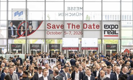 A large crowd of people attends the real estate trade fair EXPO REAL in the entrance hall of Messe München. Above the visitors hangs a large banner reading “Save the Date – October 5–7, 2026” along with the EXPO REAL logo. In the background, there are information booths, digital display screens, and glass facades letting in daylight.