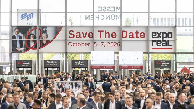 Eine große Menschenmenge besucht die Immobilienmesse EXPO REAL im Eingangsbereich der Messe München. Über den Köpfen der Besucher hängt ein großes Banner mit der Aufschrift „Save the Date – October 5–7, 2026“ und dem Logo der Messe EXPO REAL. Im Hintergrund sind Informationsstände, digitale Anzeigetafeln und Glasfassaden mit Tageslicht zu sehen.