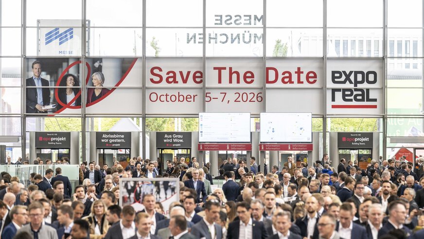A large crowd of people attends the real estate trade fair EXPO REAL in the entrance hall of Messe München. Above the visitors hangs a large banner reading “Save the Date – October 5–7, 2026” along with the EXPO REAL logo. In the background, there are information booths, digital display screens, and glass facades letting in daylight.