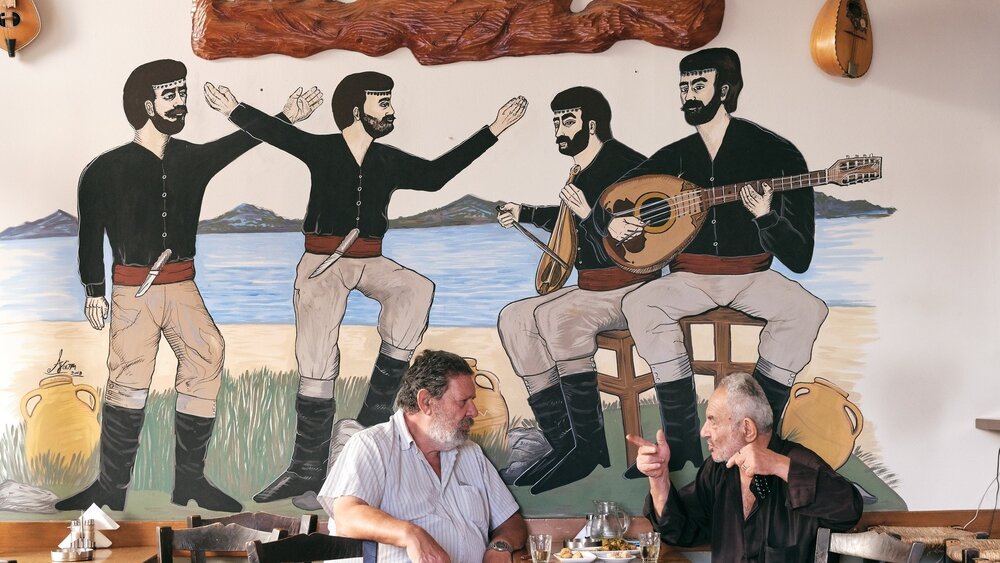 Zwei Männer unterhalten sich an einem Tisch vor einem Wandgemälde, das Musiker und Tänzer in traditioneller Kleidung zeigt.