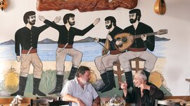 Zwei Männer unterhalten sich an einem Tisch vor einem Wandgemälde, das Musiker und Tänzer in traditioneller Kleidung zeigt.