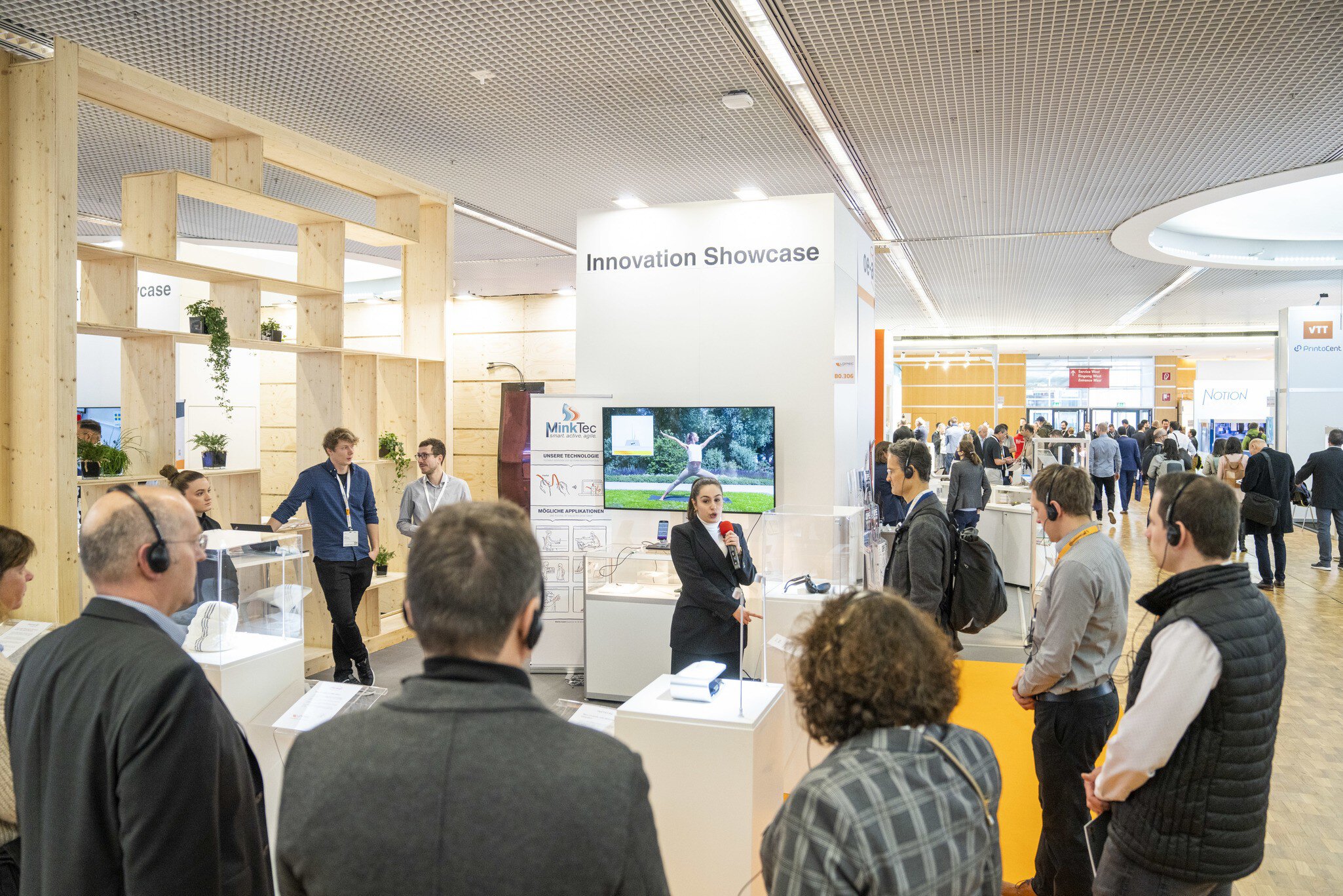 A woman with a microphone stands at a trade fair stand while a group of trade fair visitors wearing headphones listen to her with interest.