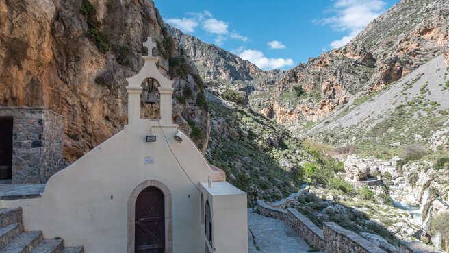 Kleine weiße Kapelle, die in einem Gebirgstal unter teilweise bewölktem Himmel in einen felsigen Abhang gebaut wurde.