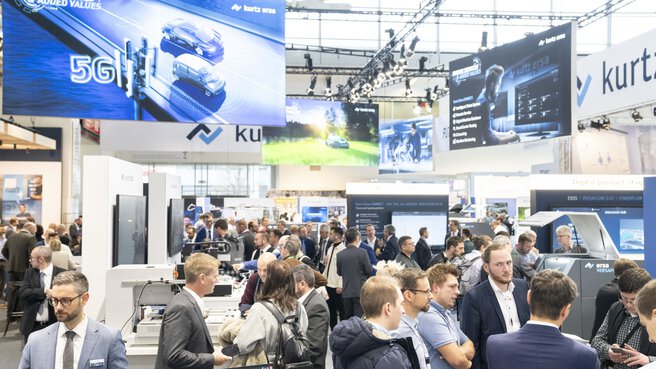 Blick in eine geschäftige Ausstellungshalle der productronica mit vielen Ständen und zahlreichen Werbebannern an der Decke.