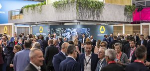 Großer Messestand von „Commerz Real“ auf einer gut besuchten Fachmesse, mit türkisfarbenem Banner, zweistöckigem Aufbau, moderner Architektur, Pflanzen auf der oberen Ebene und vielen Besuchern in Businesskleidung im Gespräch.