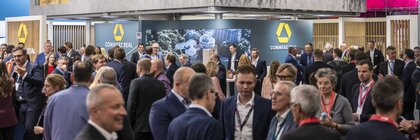 Large “Commerz Real” exhibition booth at a busy trade fair, featuring a turquoise banner, a two-level modern structure, greenery on the upper level, and many attendees in business attire engaged in conversations.
