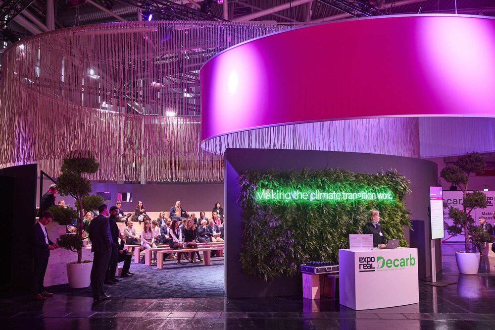 Exhibition hall: a green wall with neon lettering reading: ‘Making the climate transition work’. In front of it, a woman is standing at a counter.