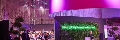 Eine Messehalle: Eine begrünte Wand mit Neonschriftzug: „Making the climate transition work“. Davor steht eine Frau an einem Tresen mit Logos von EXPO REAL und Decarb.