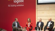 Fünf Personen sitzen auf der Bühne der transport logistic vor Publikum. Eine Frau im roten Anzug spricht gerade und gestikuliert mit beiden Händen.