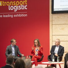 Fünf Personen sitzen auf der Bühne der transport logistic vor Publikum. Eine Frau im roten Anzug spricht gerade und gestikuliert mit beiden Händen.