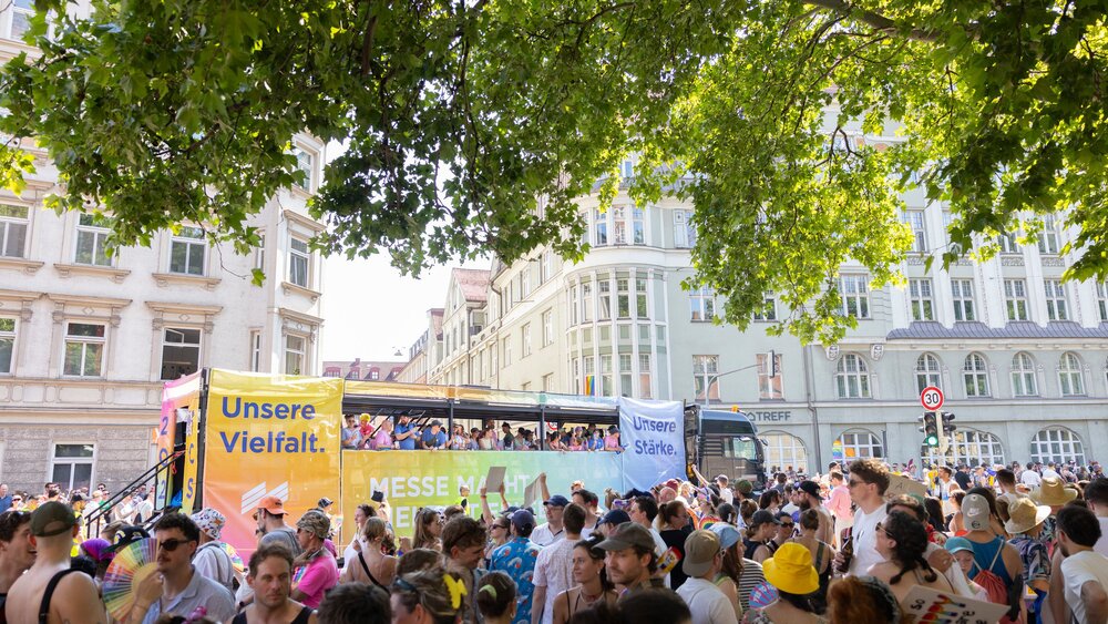 A lively street parade featuring a colorful bus and a large crowd, with banners reading “Our Diversity” and “Our Strength.”