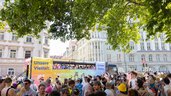 Eine lebhafte Parade auf der Straße mit einem bunten Bus und einer großen Menschenmenge, mit Transparenten mit den Aufschriften „Unsere Vielfalt“ und „Unsere Stärke“.