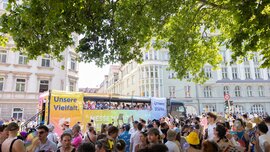 A lively street parade featuring a colorful bus and a large crowd, with banners reading “Our Diversity” and “Our Strength.”