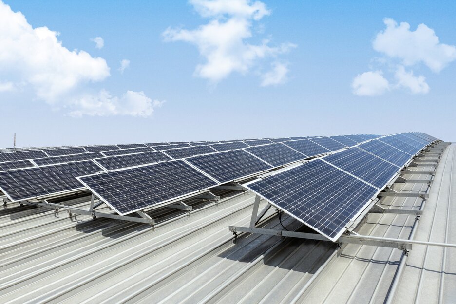Ein gewelltes Metalldach mit zahlreichen Solarpanelen und einem blauen Himmel mit ein paar weißen Wolken darüber.